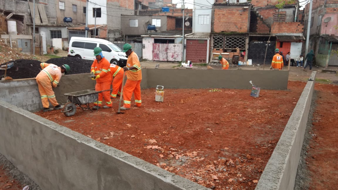 #PraCegoVer - Trabalhadores da Subprefeitura finalizam mureta de tijolos na praça que está sendo revitalizada. Ao fundo, casas do local.