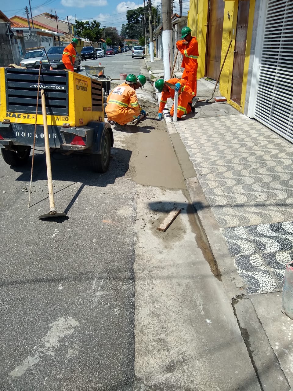 #PraCegoVer - Trabalhadores da Subprefeitura finalizam sarjeta na via. A calçada é feita de ladrilhos imitando pedras portuguesas. Há um compressor amarelo à esquerda.