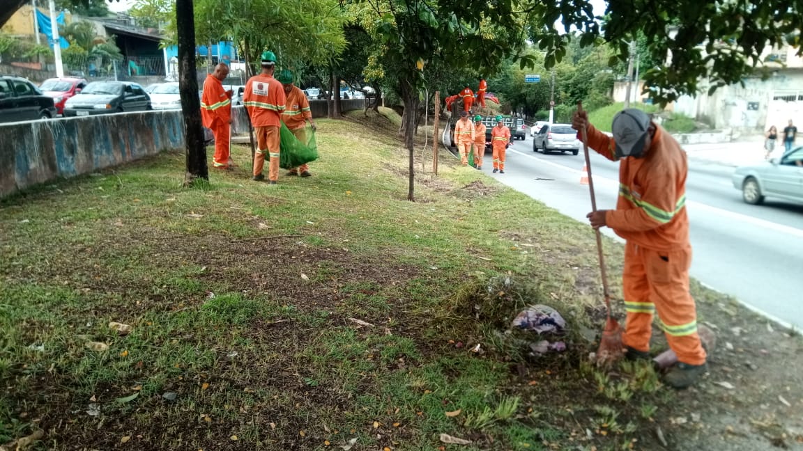 #PraCegoVer - Trabalhadores da Subprefeitura cortam grama e recolhem o material cortado no canteiro central da via. Há árvores. E uma defensa de concreto do lado esquerdo.