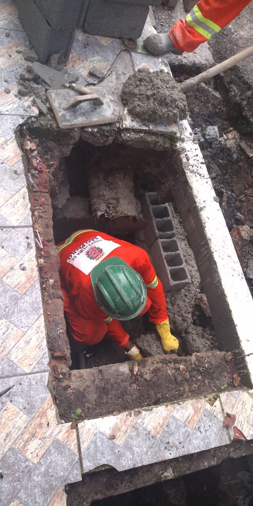 #PraCegoVer - Trabalhador da Subprefeitura está dentro da boca de lobo, assentando blocos de concreto. Um outro prepara massa de cimento.