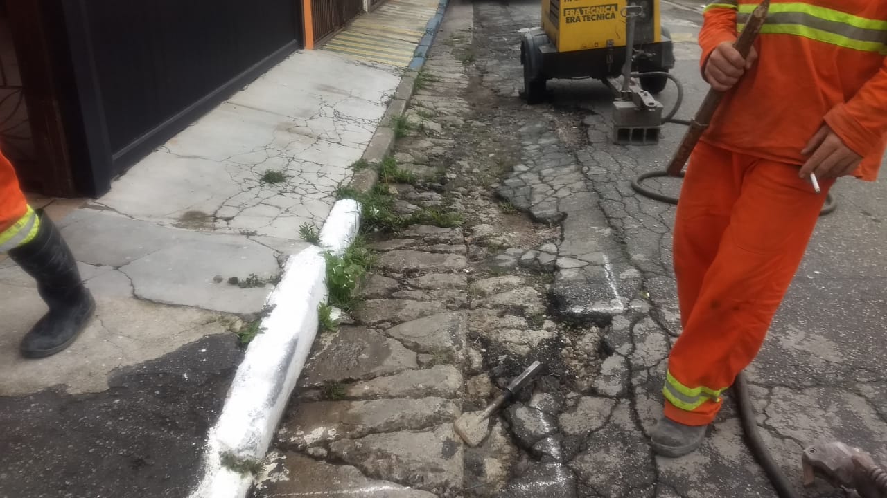 #PraCegoVer - Trabalhadores da Subprefeitura começam a escavar a rua para retirada da antiga sarjeta. Ao fundo, um aparelho que aciona a britadeira.