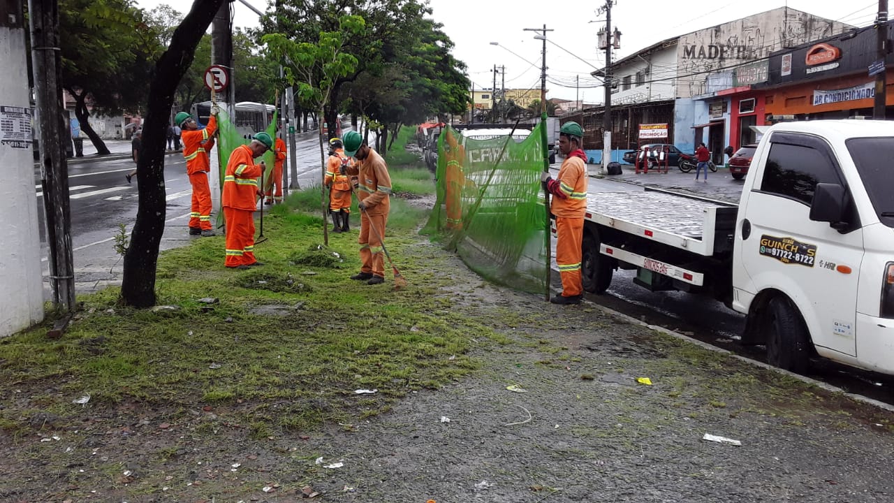 #PraCegoVer - Trabalhadores da Subprefeitura cortam grama no canteiro central da via. Outros seguram proteção de tela.