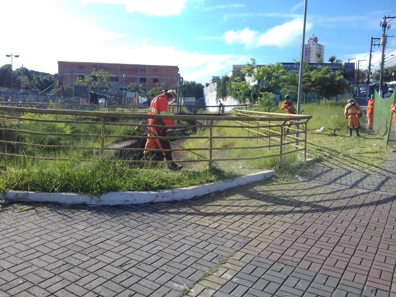 #PraCegoVer - Trabalhadores da Subprefeitura cortam grama em torno de um córrego, protegido por guarda-corpo. Outros seguram proteção de tela.