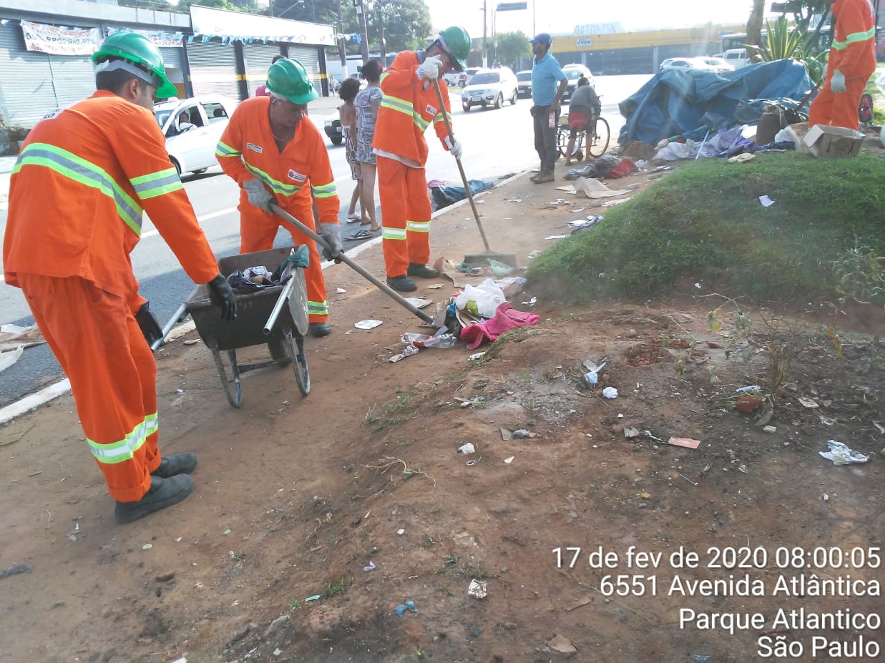 #PraCegoVer - Trabalhadores da Subprefeitura recolhem lixo no canteiro central da via. Um dos trabalhadores maneja um carrinho de mão; outros dois usam vassouras. Ao fundo, há uma tenda azul.