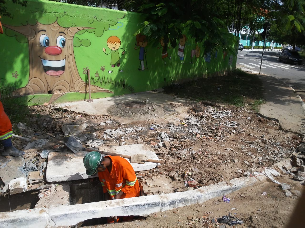 #PraCegoVer - Trabalhador da Subprefeitura está dentro de uma boca de lobo, escavando. O passeio está parcialmente destruído. Ao fundo, em um muro possivelmente de escola, há um desenho de uma árvore e um menino.