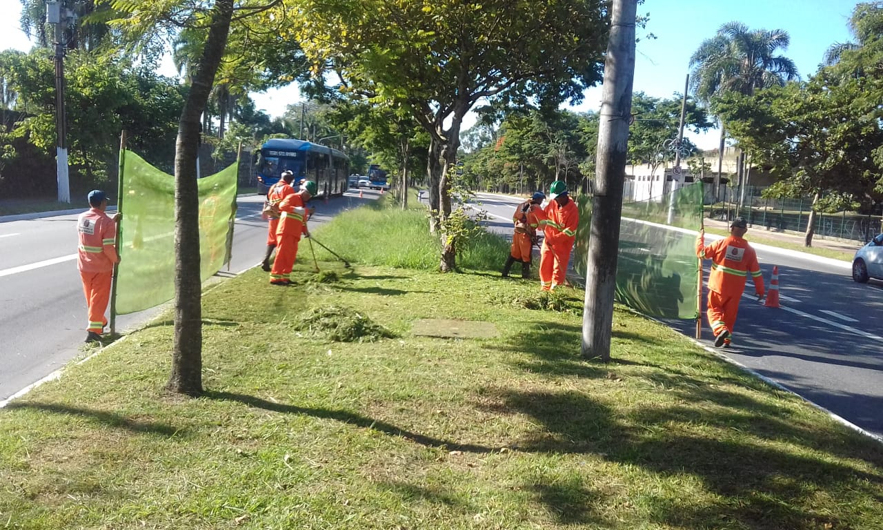 #PraCegoVer - Trabalhadores da Subprefeitura cortam grama no canteiro central da via. Outros seguram proteções de tela.