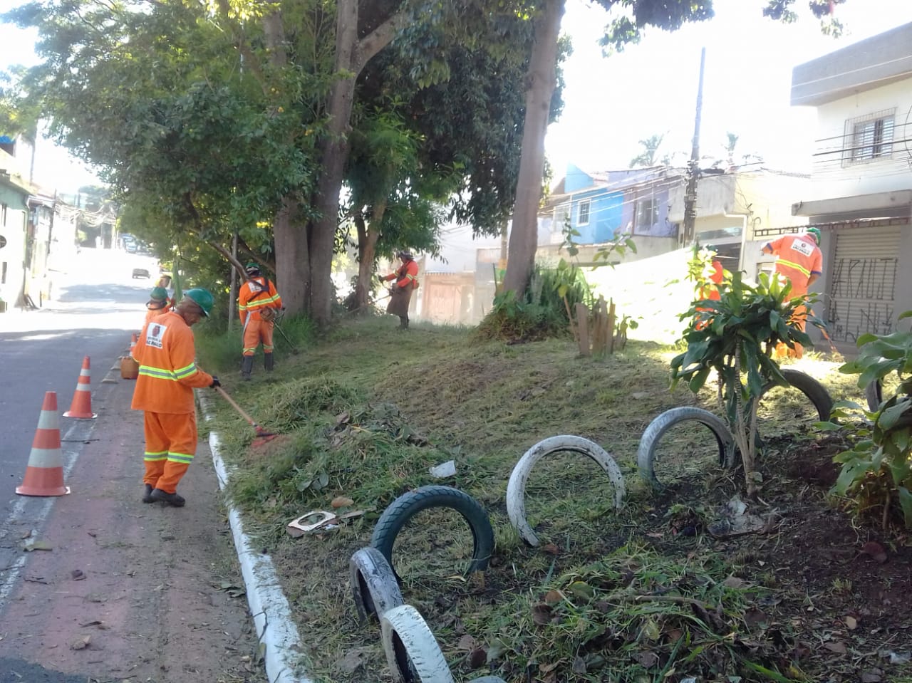 #PraCegoVer - Trabalhadores da Subprefeitura cortam grama no canteiro central da via. Há árvores ao fundo. No primeiro, plano, uma cercadura de canteiro feita com pneus velhos.