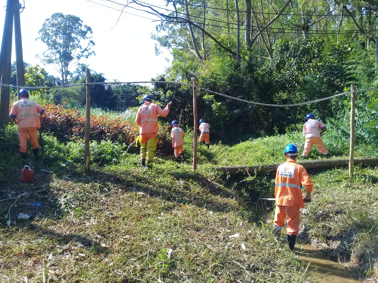 #PraCegoVer - Trabalhadores da Subprefeitura limpam o córrego. Há farta vegetação em volta.