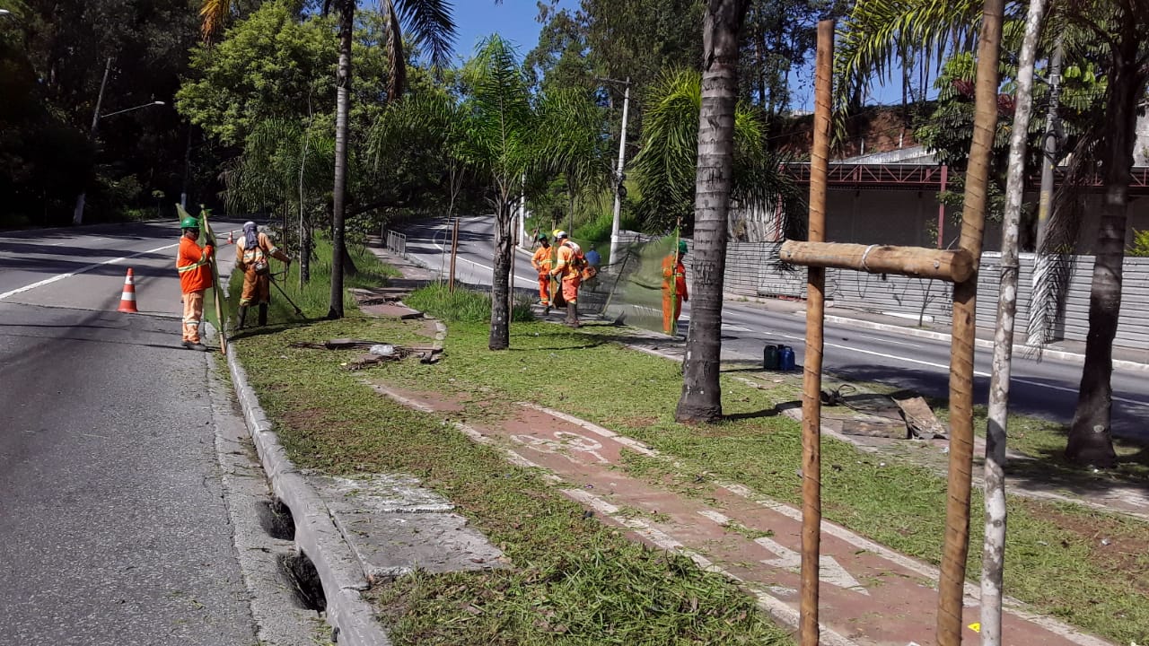 #PraCegoVer - Trabalhadores da Subprefeitura cortam grama no canteiro central da via. No canteiro, há uma ciclovia com duas pistas.