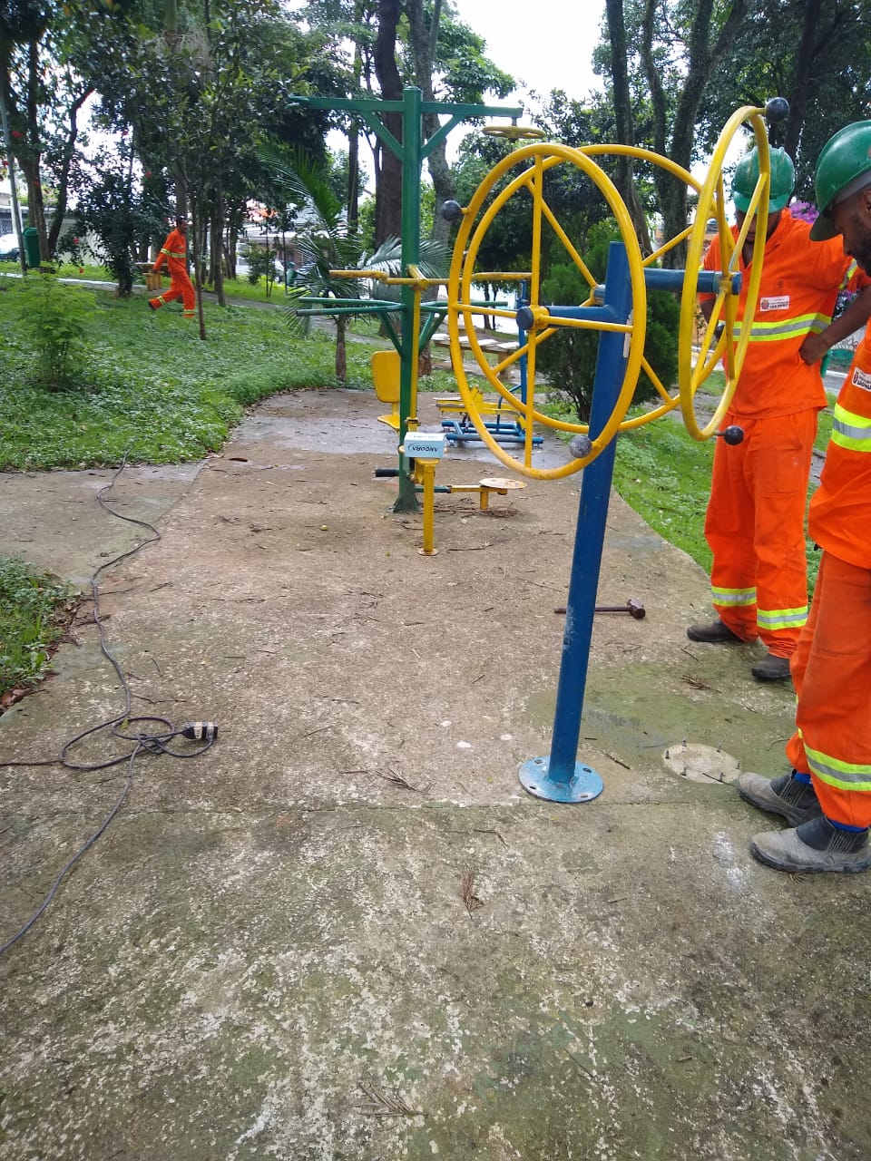 #PraCegoVer - Trabalhadores da Subprefeitura finalizam instalação de aparelhos de ginástica na calçada da praça. Há gramados e árvores no local.