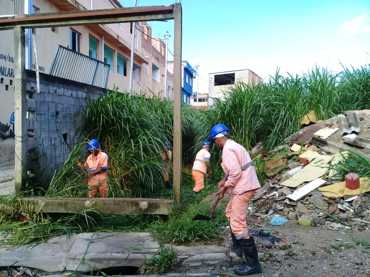 #PraCegoVer - Trabalhadores da Subprefeitura limpam o córrego. De lado esquerdo, há uma passagem e um pequeno prédio. Do lado direito, um amontado de entulho e lixo. Sobre o córrego, há uma armação em concreto.