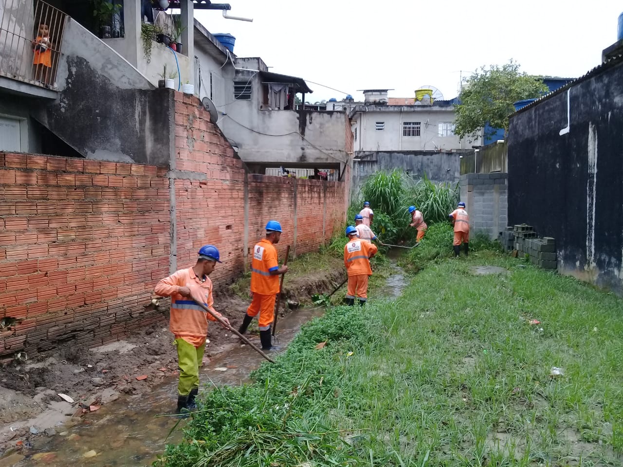 #PraCegoVer - Trabalhadores da Subprefeitura limpam córrego ao lado de uma casa com muro de tijolos, do lado esquerdo. Há outra construção do lado direito.