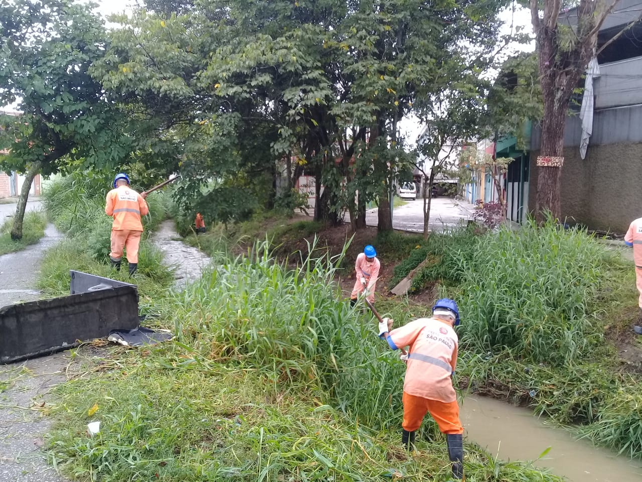 #PraCegoVer - Trabalhadores da Subprefeitura fazem limpeza no córrego, ao lado de algumas construções. Há árvores ao fundo.