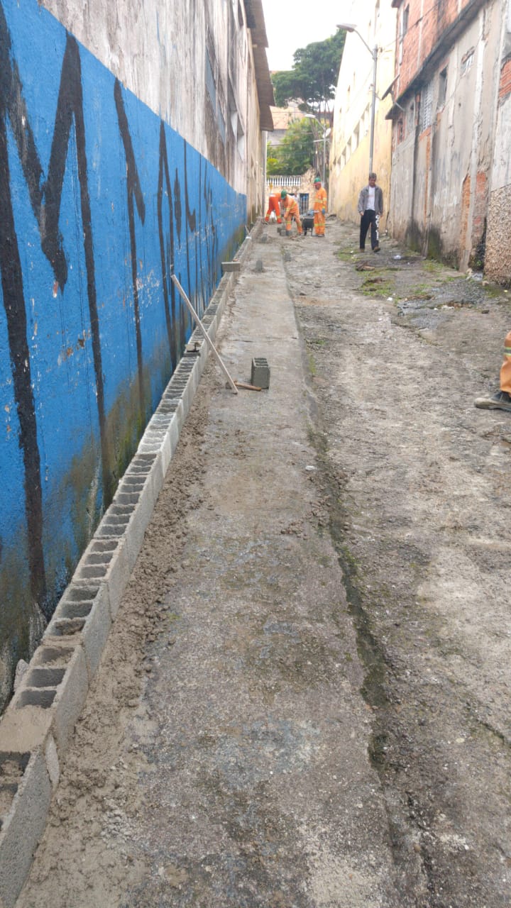 #PraCegoVer - Trabalhadores da Subprefeitura reformam o piso da viela. Do lado esquerdo, há uma fileira de blocos de concreto, apoiados em um muro azul marcado por pichações.