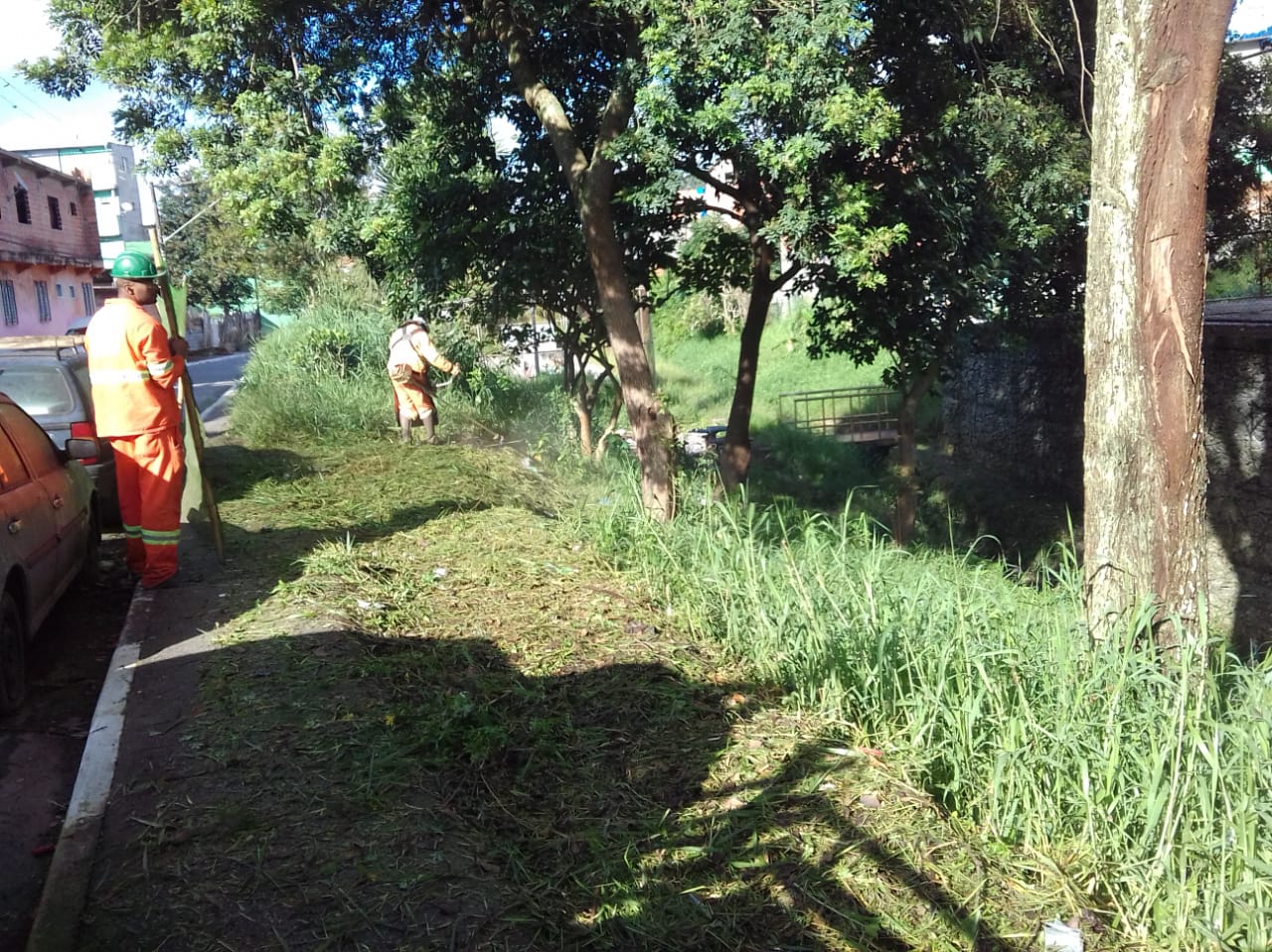 #PraCegoVer - Trabalhadores da Subprefeitura cortam grama e mato à beira de córrego. Há árvores no local.