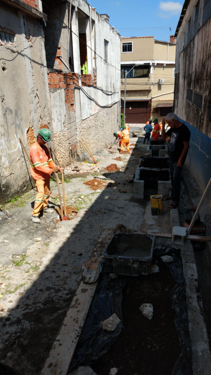 #PraCegoVer - Trabalhadores da Subprefeitura instalam floreiras na viela. O local é cercado por construções cujas paredes estão sendo rebocadas.