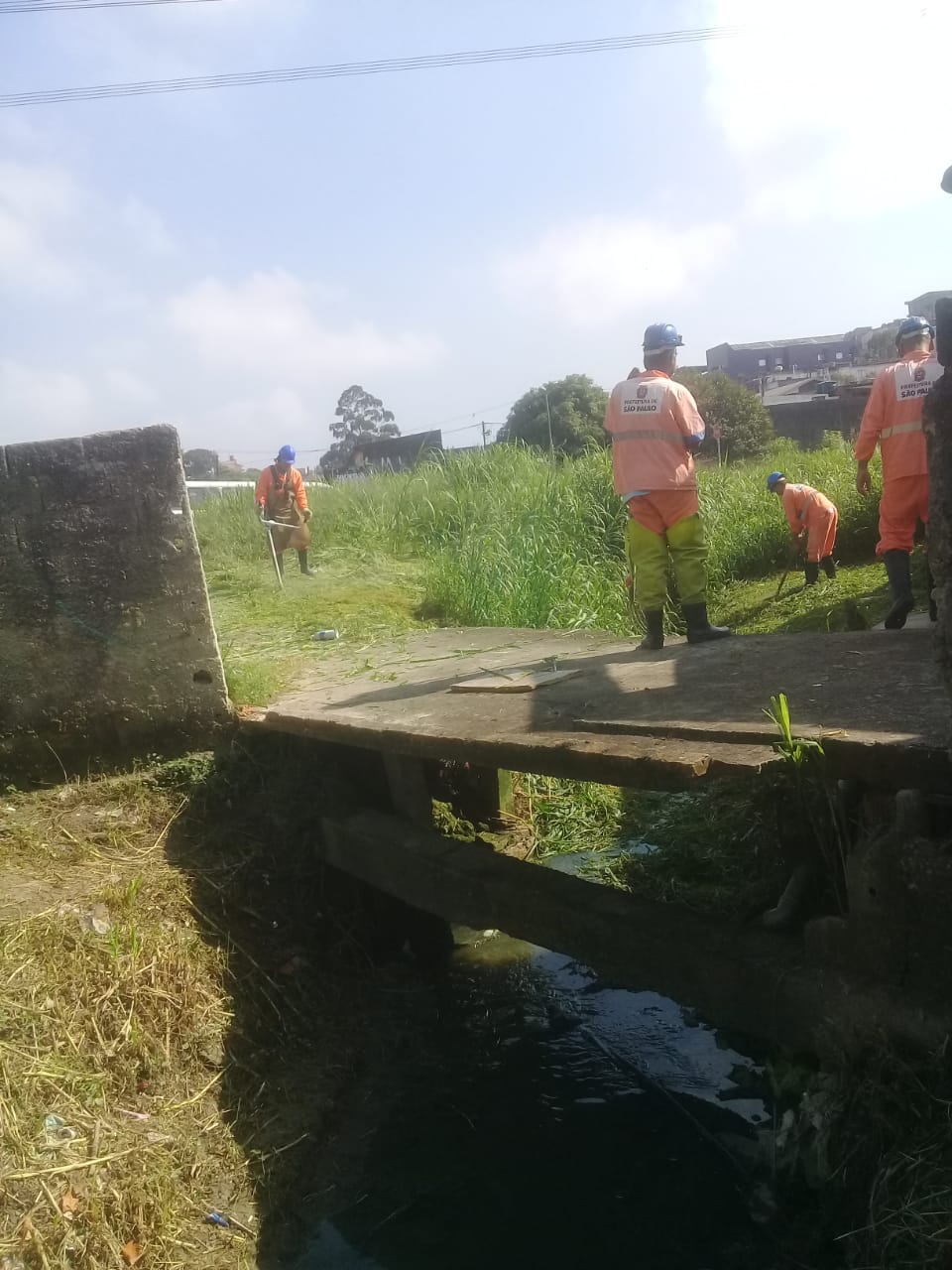#PraCegoVer - Trabalhadores da Subprefeitura cortam mato e grama ao lado de uma ponte de concreto sobre o córrego.