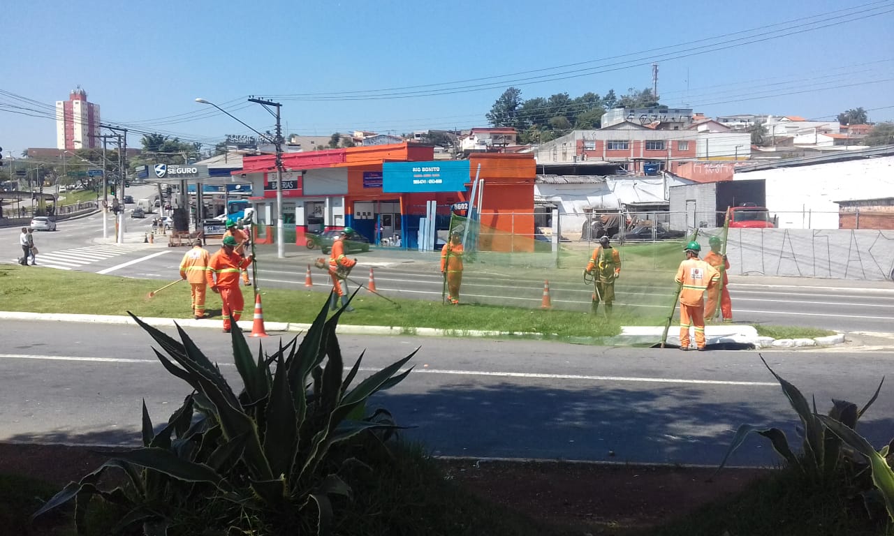#PraCegoVer - Trabalhadores da Subprefeitura cortam grama no canteiro central que liga as vias. Outros seguram proteção de tela.