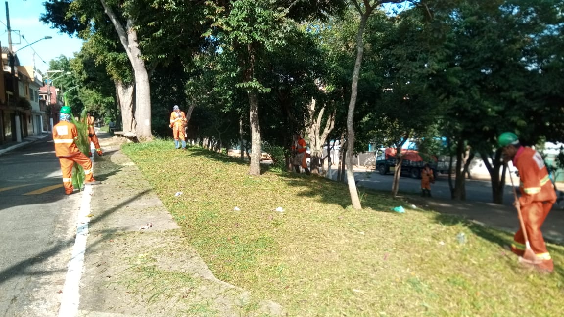 #PraCegoVer - Trabalhadores da Subprefeitura Cortam grama entre árvores. Outros trabalhadores seguram a proteção de tela.