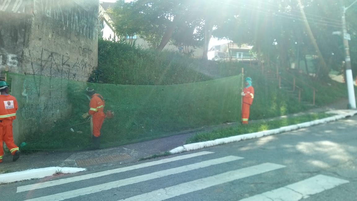 #PraCegoVer - Trabalhadores da Subprefeitura cortam grama no encontro de duas ruas. Outros seguram a proteção de tela.