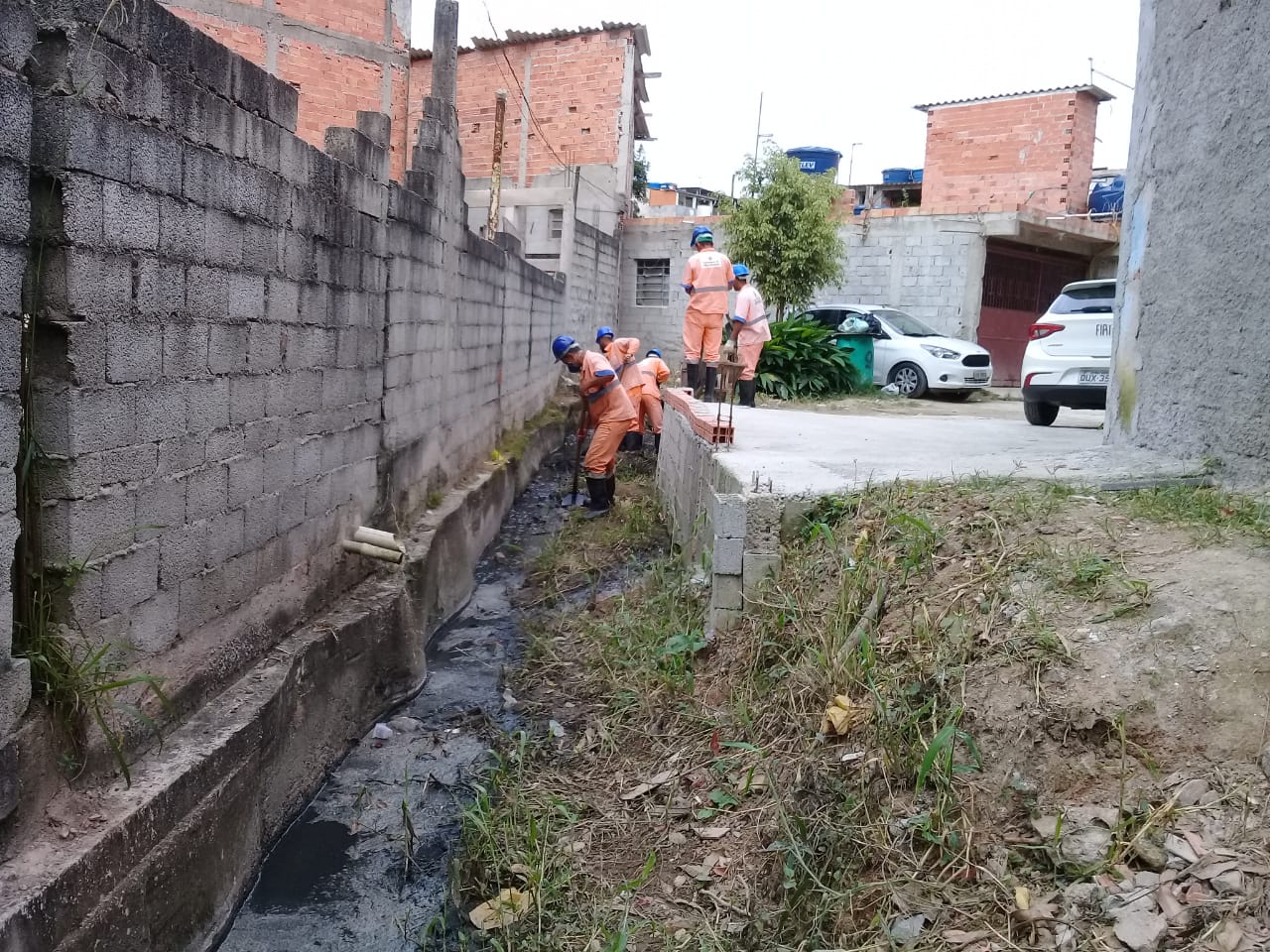 #PraCegoVer - Trabalhadores da Subprefeitura removem obstruções do córrego. Do lado esquerdo, há um muro de blocos de concreto. Do lado direito, uma plataforma de cimento.