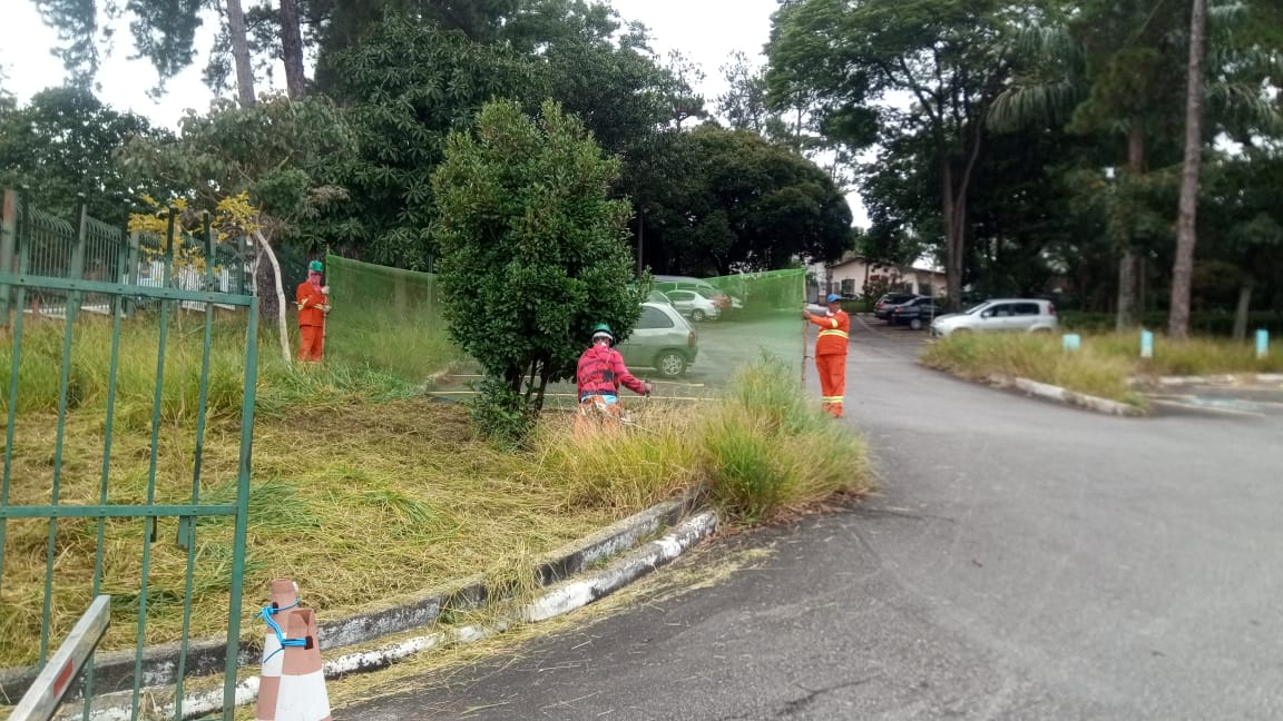 #PraCego Ver - Trabalhadores da Subprefeitura cortam grama nos jardins da Subprefeitura. Há árvores no primeiro plano e ao fundo. À esquerda, o portão de acesso à Subprefeitura.