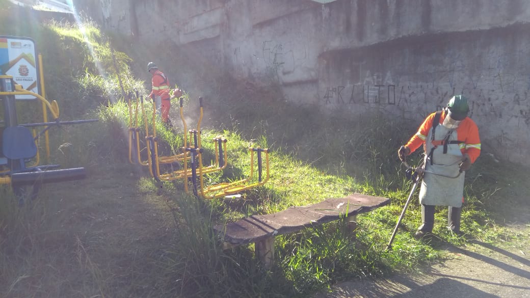 #PraCegoVer - Trabalhadores da Subprefeitura cortam grama em local com aparelhos para Terceira Idade (ATI). Um radio de sol perpassa toda a cena.