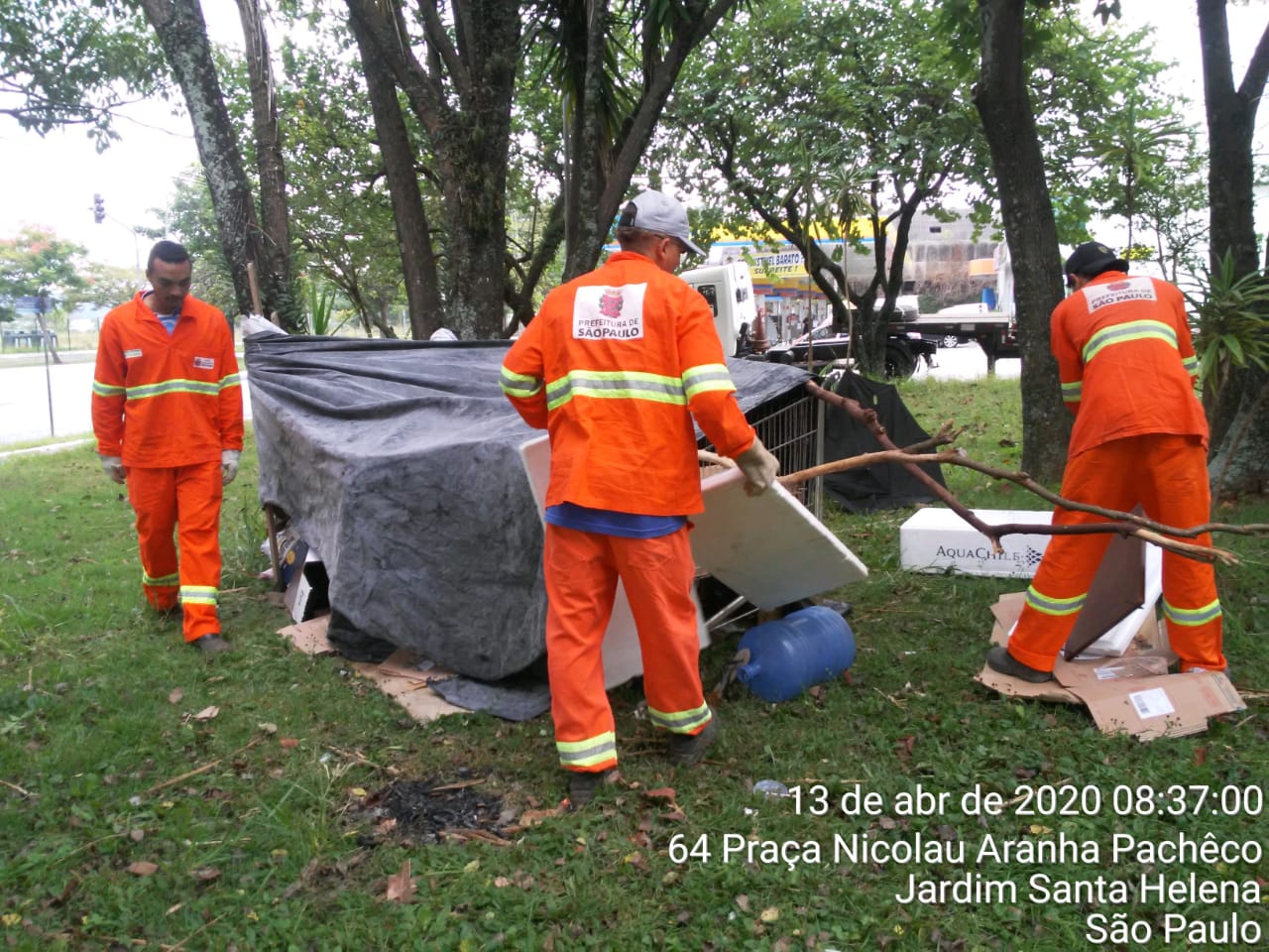 #PraCegoVer - Trabalhadores da Subprefeitura removem barracas da praça. Há árvores. O local é todo gramado.