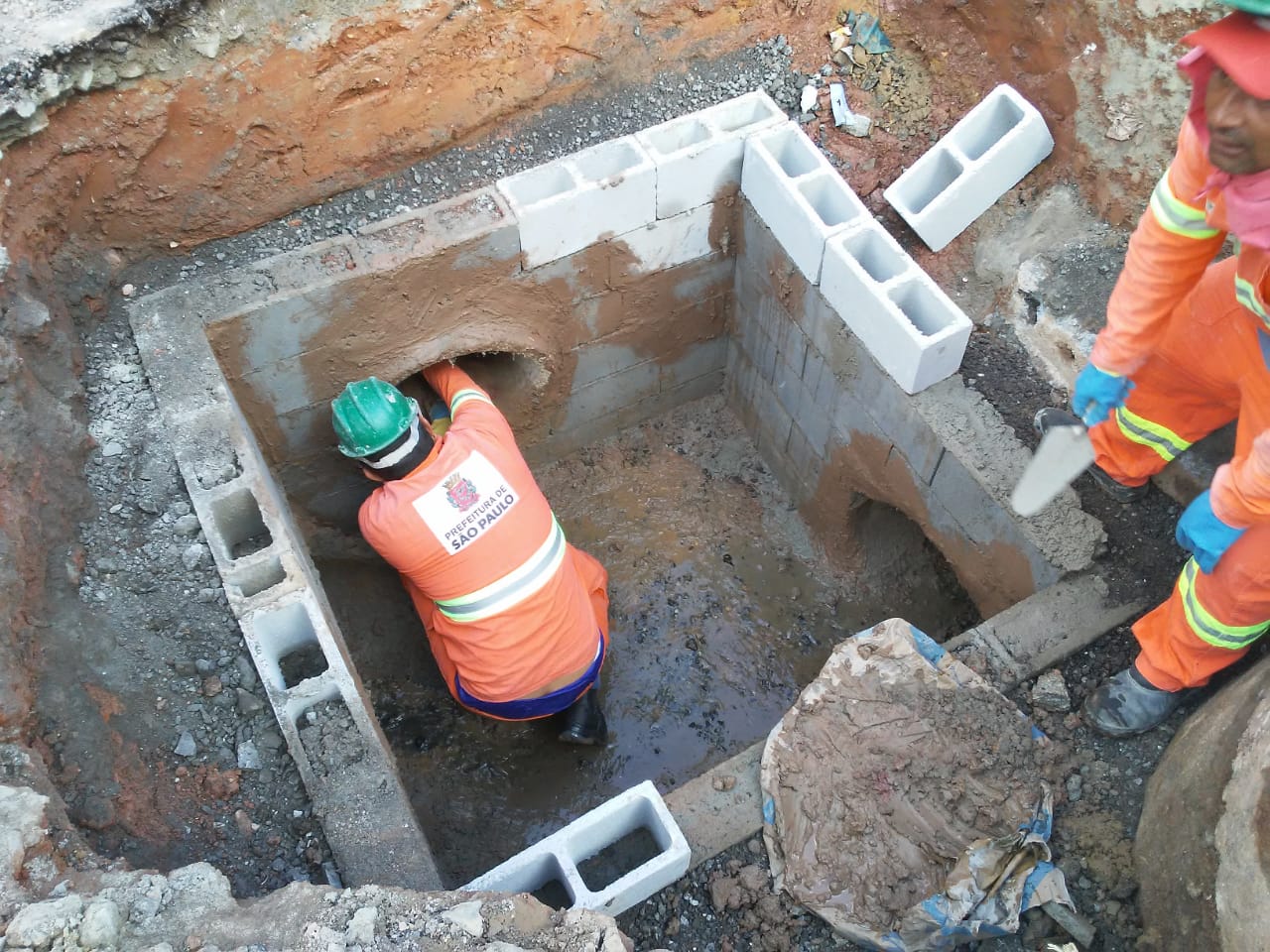 #PraCegoVer - Trabalhador da Subprefeitura constrói caixa de blocos de concreto. Na galeria, desembocam pelo menos dois tubos.