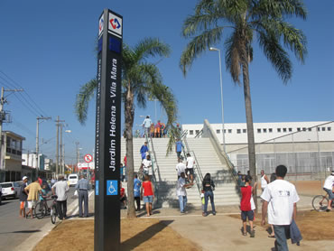 A foto feita em plano aberto mostra a entrada da estação Jardim Helena, em foto diversas pessoas estão em frente a estação.