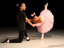 A imagem apresenta duas crianças dançando, uma ajoelhada de preto e a outra de rosa, com um dos pés esticados, em uma possível apresentação de balé.