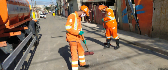 Dois funcionários da prefeitura com uniformes laranjas segurando vassouras limpando a rua