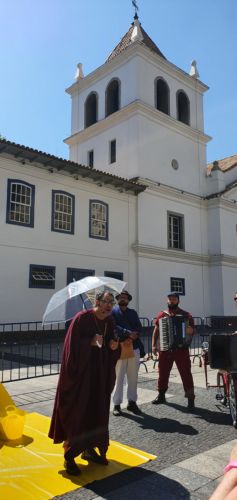 Ator com túnica vinho e guarda-chuva transparente interpreta o Padre Anchieta e fala da 1]a construção que daria a criação da cidade de São Paulo
