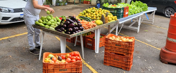 No primeiro plano da foto aparecem caixas de frutas e legumes no estacionamento da Subprefeitura Itaim Paulista. No segundo plano da foto aparece a banca comunitária com frutas, legumes e hortaliças.