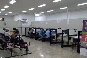 Imagem da praça de atendimento da Subprefeitura Santo Amaro com balcões numerados para o atendimento à esquerda e à direita estão cadeiras com munícipes sentados aguardando serem atendidos