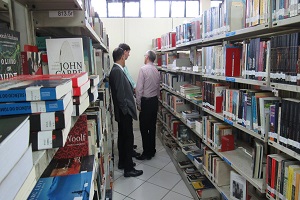 A foto apresenta três pessoas em um corredor de uma biblioteca municipal. Ao lado, prateleiras com livros dos dois lados