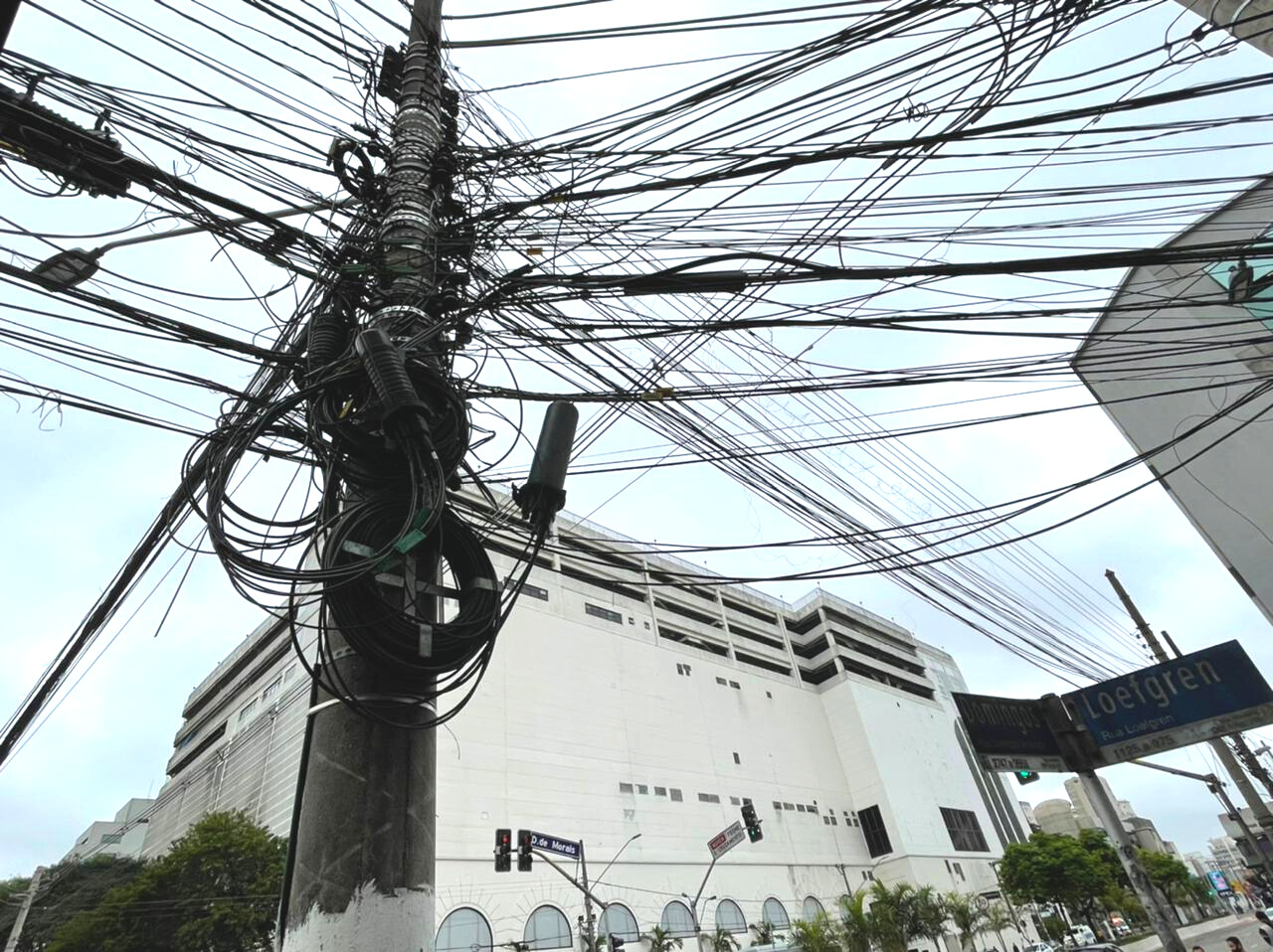 Imagem mostra fios emaranhados em um poste com o Shopping santa Cruz ao fundo. No canto direito há a placa da esquina da Rua Loefgreen e da Avenida Domingos de Morais.