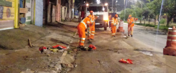 Quatro funcionários da Prefeitura, com uniformes laranja, trabalham na limpeza de lama em uma rua, à frente de um caminhão, com faróis acesos e cones de isolamento na lateral direita.