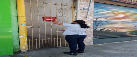 Funcionária da Ecourbis usando uniforme de calça preta e camisa azul, está de costas e de  frente para o portão de uma moradia depositando o folheto na caixinha vermelha que está pendurada no portão para o depósito de cartas.
