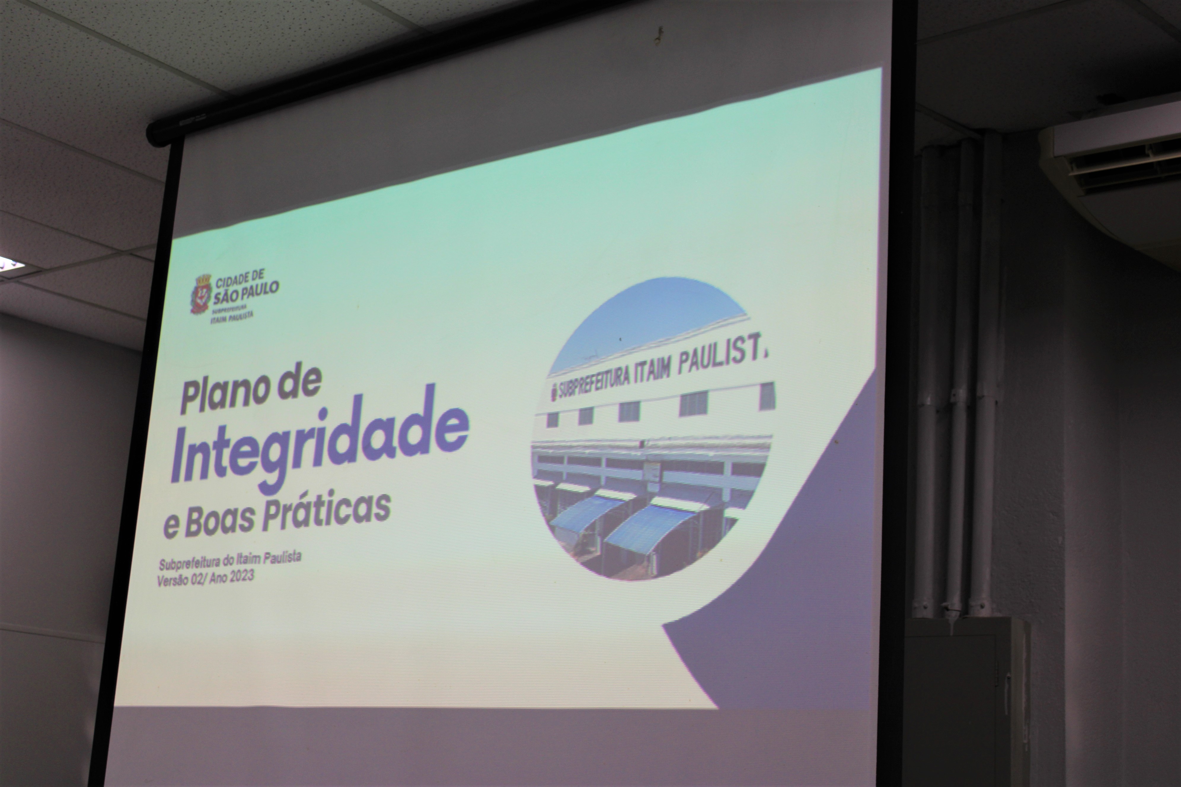 Uma foto de um telão com o primeiro slide da apresentação do Plano de Integridade e Boas Práticas. Na imagem, há os dizeres: Plano de Integridade e Boas Práticas, subprefeitura do Itaim Paulista, versão 02/Ano 2023.
