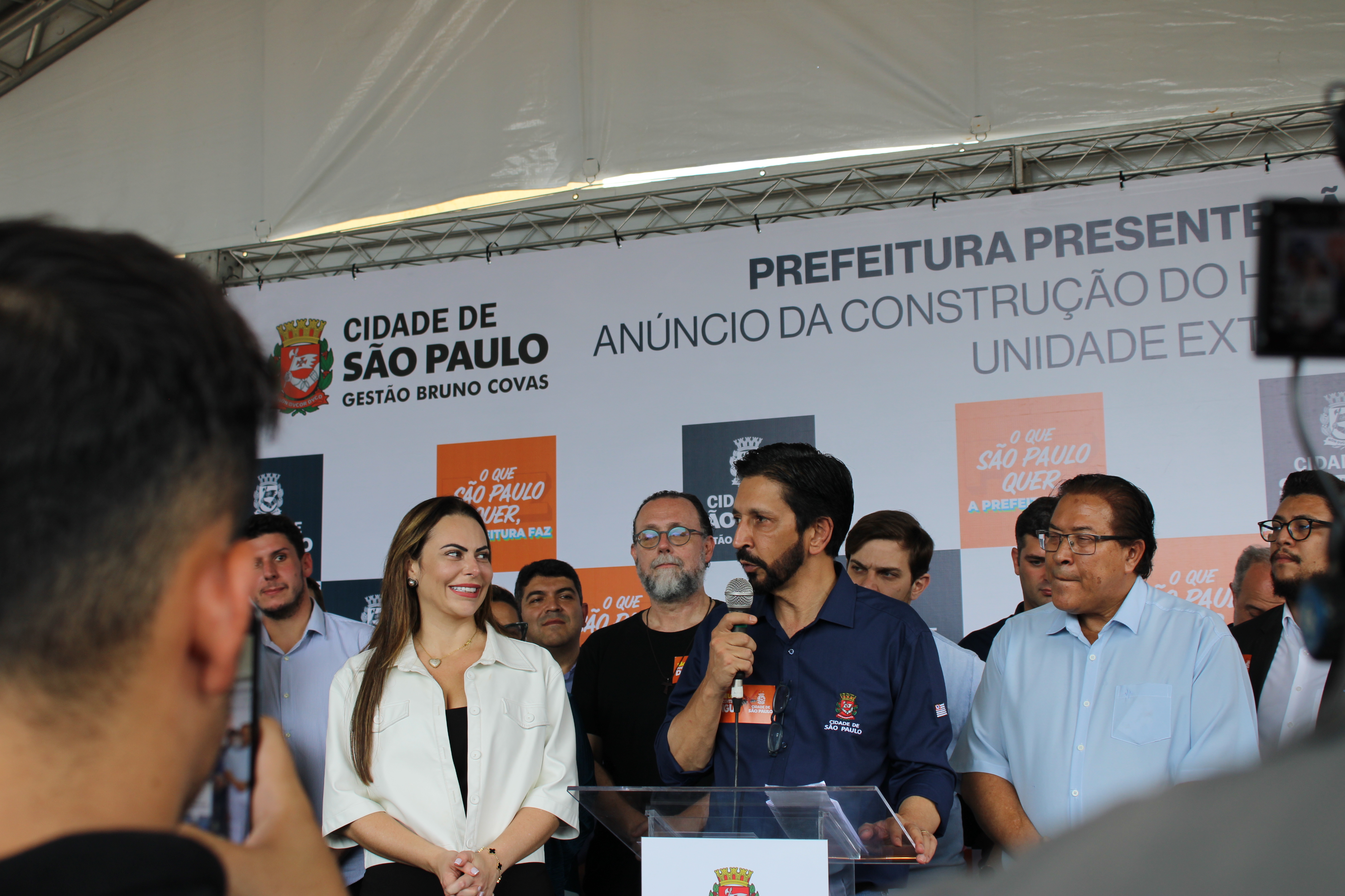 Foto do prefeito Ricardo Nunes falando ao microfone ao lado de sua esposa, a primeira dama Regina Carnovale, que está olhando e sorrindo para ele, enquanto outros membros da equipe de governo estão atrás dele no palanque.
