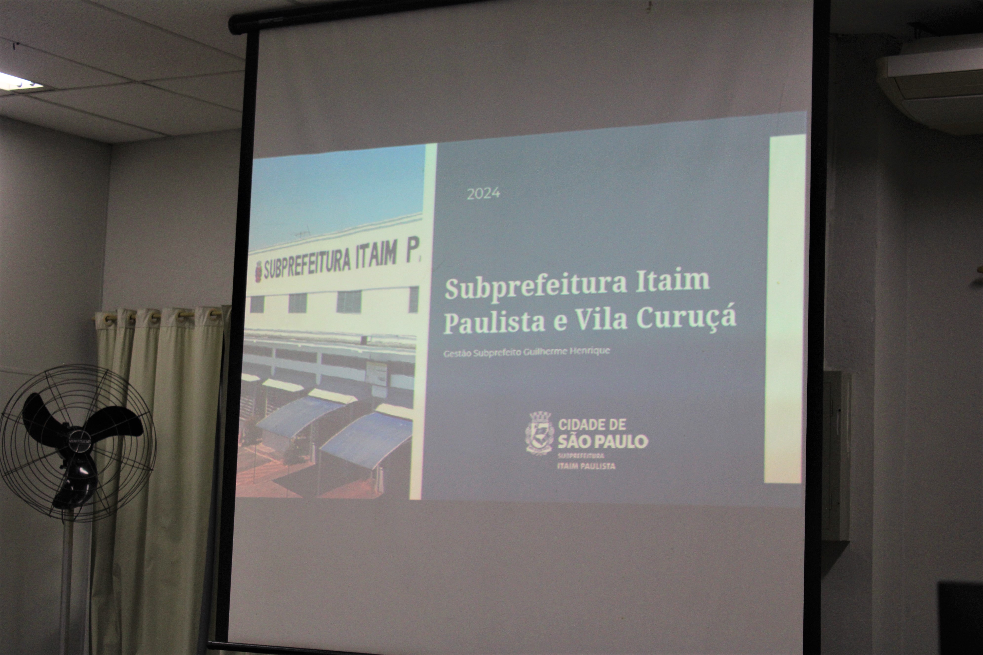 Foto de uma apresentação em um telão com os escritos: Subprefeitura Itaim Paulista e Vila Curuçá. Gestão do Subprefeito Guilherme Henrique.