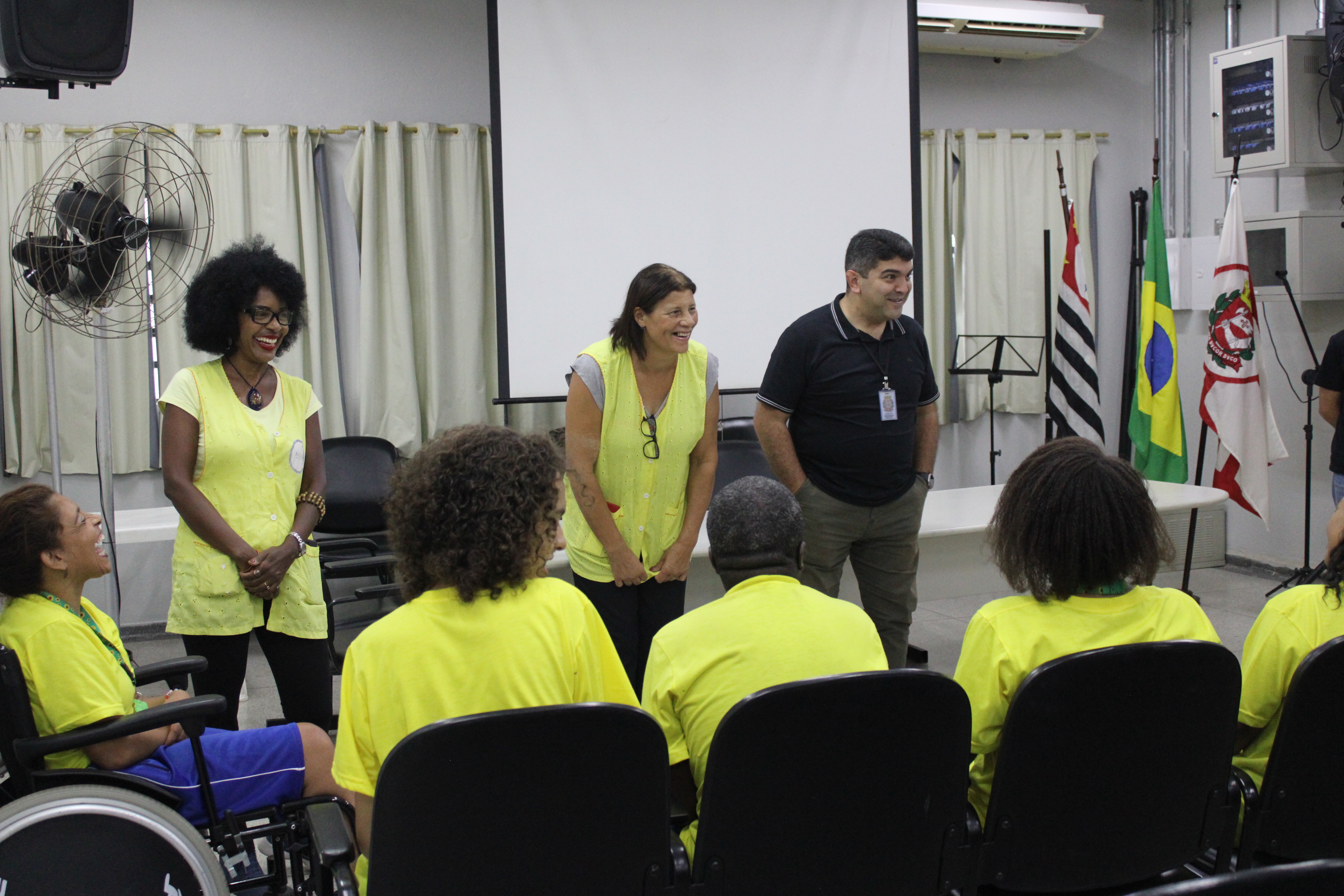 Foto de alunos da Associação AECOM de camiseta verde sentados e olhando o subprefeito sorrindo ao lado das responsáveis pela entidade que cuida de pessoas com deficiência.