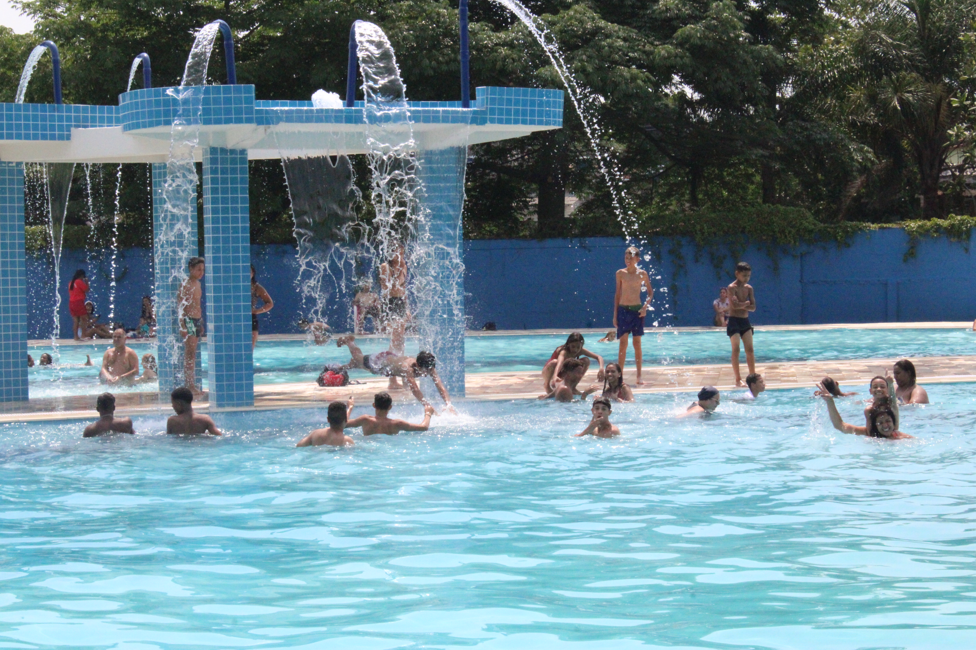 Uma imagem ao ar livre de com várias crianças e adultos dentro de uma piscina azul grande com uma espécie de chafariz caindo água