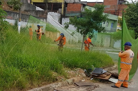 Cinco funcionários vestidos com macacão cor laranja sendo três cortando mato com equipamento do tipo roçadeira enquanto os outros dois funcionários seguram uma rede de proteção de cor verde para que o mato cortado não se espalhe