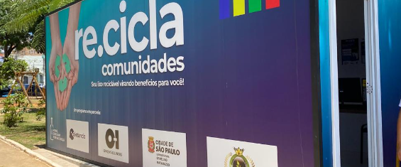 A imagem mostra um contêiner de reciclagem com uma grande faixa publicitária em sua lateral. A faixa tem um fundo que varia de azul claro na parte superior a um roxo mais escuro na parte inferior. No centro superior, em letras brancas grandes, está escrito 