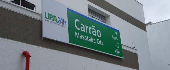 Placa com os dizeres UPA 24h - Carrão Massataka Ota -pintada em fundo branco e verde afixada na parede do prédio da Unidade de Pronto Atendimento Carrão