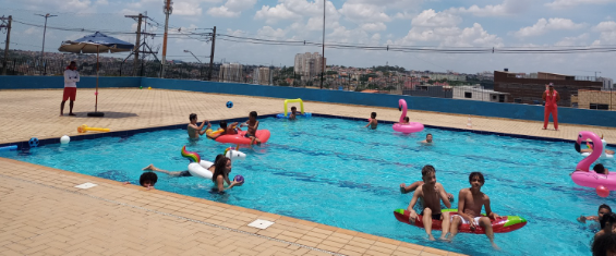 Crianças brincam em uma piscina, com boias de plástico coloridas. Um salva vidas, com uniforme laranja observa ao fundo.