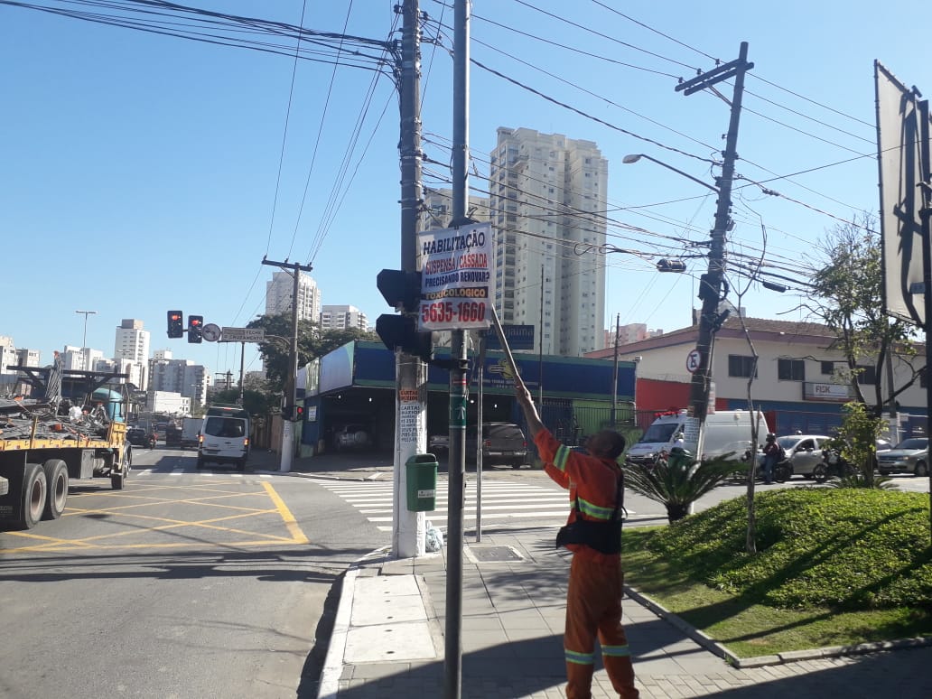 Imagem mostra funcionário vestido de laranja retirando uma faixa de um poste