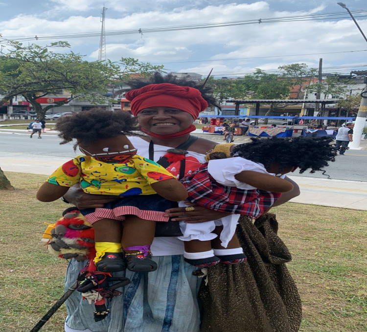 a artesã Ana Julia usando turbante vermelho com os bonecos pretos no colo o Guebo e a Mirra ela está na praça próxima das barracas de artesanato que vemos instaladas ao fundo